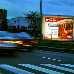 Podświetlony billboard z reklamą United Chicken przy jezdni, obok budynek i przejeżdżający samochód. Widoczny fragment przejścia dla pieszych.