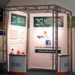 Stoisko targowe z metalową konstrukcją, banerami reklamowymi uczelni i dwoma pultami. Widoczne logo uczelni i adres strony na Facebooku. Oświetlenie punktowe.