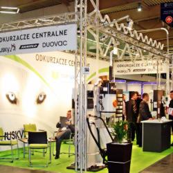 Stoisko targowe z ekspozycją odkurzaczy centralnych Husky i Duvovac, z grafiką przedstawiającą oczy. Widoczne krzesła, rośliny doniczkowe i osoby w tle.