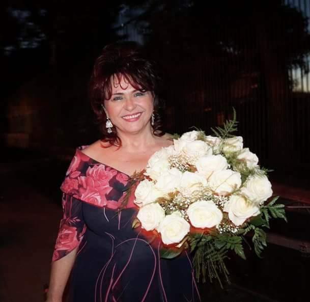 Uśmiechnięta kobieta w sukience z motywem róż trzyma bukiet białych róż, widoczne długie kolczyki.