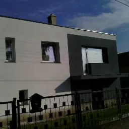 Dwukolorowa elewacja domu jednorodzinnego z płaskim dachem, widoczna zza metalowego ogrodzenia z tabliczką 'Uwaga Pies'.