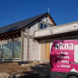 Budrop - Budowa domu jednorodzinnego w stanie surowym otwartym z widocznymi oknami i częściowo zamurowanymi otworami, na pierwszym planie firmowy samochód dostawczy z reklamą okien, drzwi, bram, rolet...