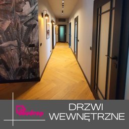 Budrop - Długi korytarz z parkietem ułożonym w jodełkę, oświetlony lampami ściennymi i sufitowymi, z trzema parami drzwi wewnętrznych z czarnymi ramami i matowym szkłem, tapeta w duże liście po lewej stronie.