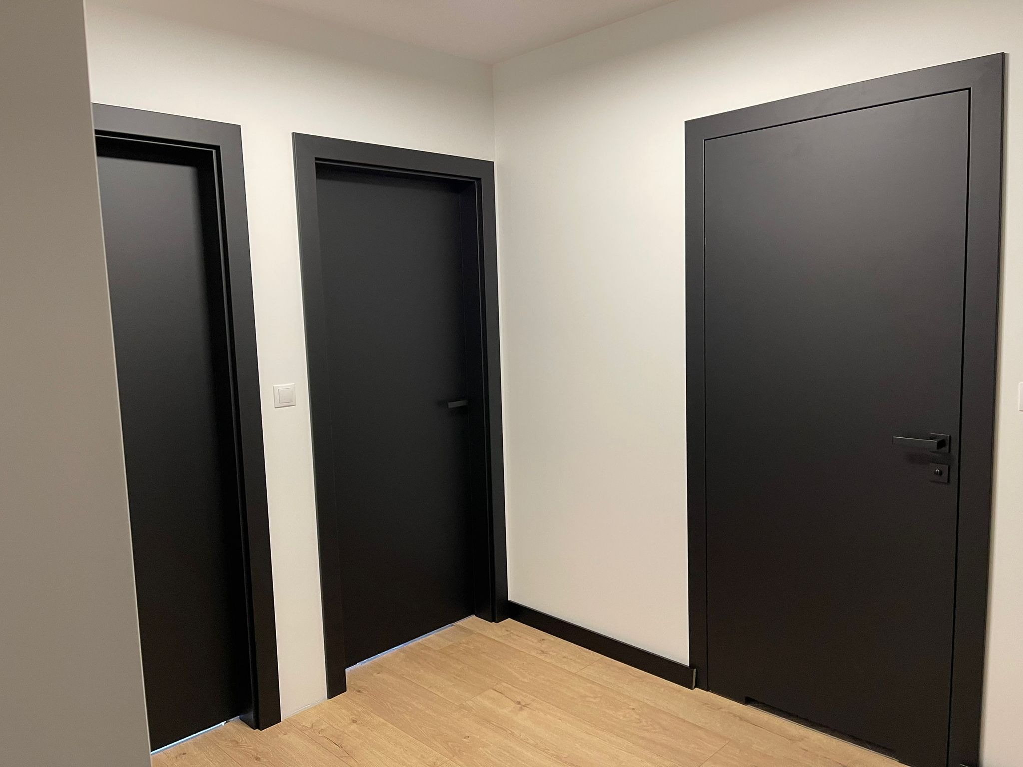 Trzy czarne, minimalistyczne drzwi wewnętrzne z czarnymi ościeżnicami i klamkami, widoczne w korytarzu z jasną podłogą i białymi ścianami.