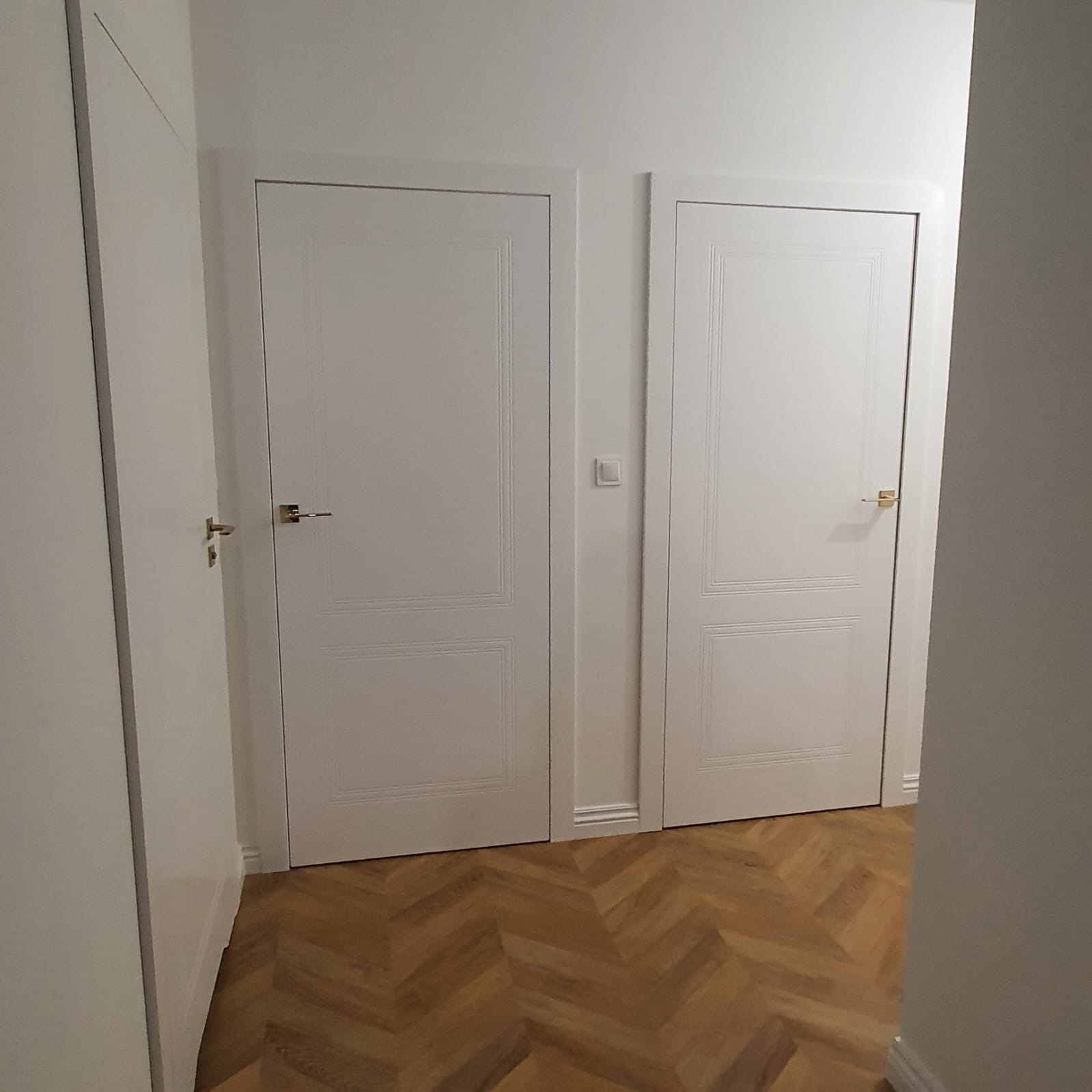 Dwa białe, klasyczne drzwi wewnętrzne z delikatnym frezowaniem, zamontowane w białej ścianie na tle parkietu ułożonego w jodełkę.