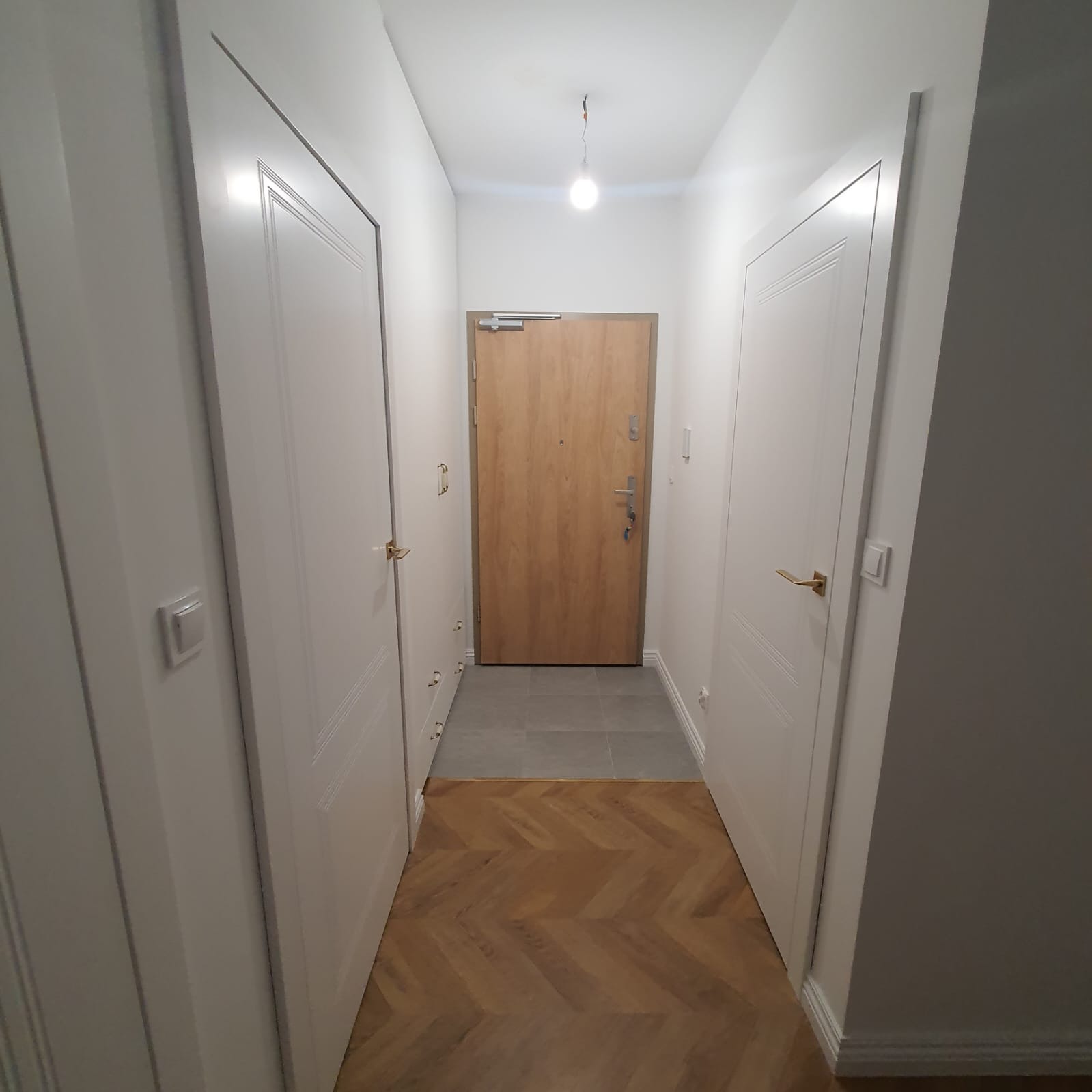 Korytarz z drewnianą podłogą ułożoną w jodełkę, białymi drzwiami wewnętrznymi po bokach i drewnianymi drzwiami wejściowymi na końcu, oświetlony pojedynczą żarówką.
