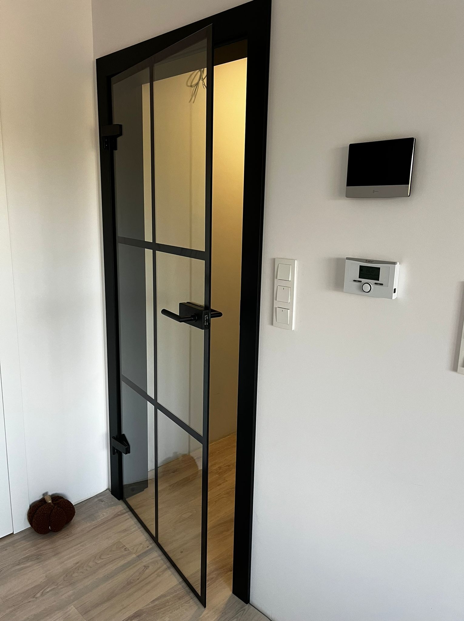 Otwarta, nowoczesna, szklana brama z czarną ramą i uchwytem, widok na wnętrze pomieszczenia, obok dekoracyjna dynia na podłodze, na ścianie włączniki i panel sterowania ogrzewaniem.