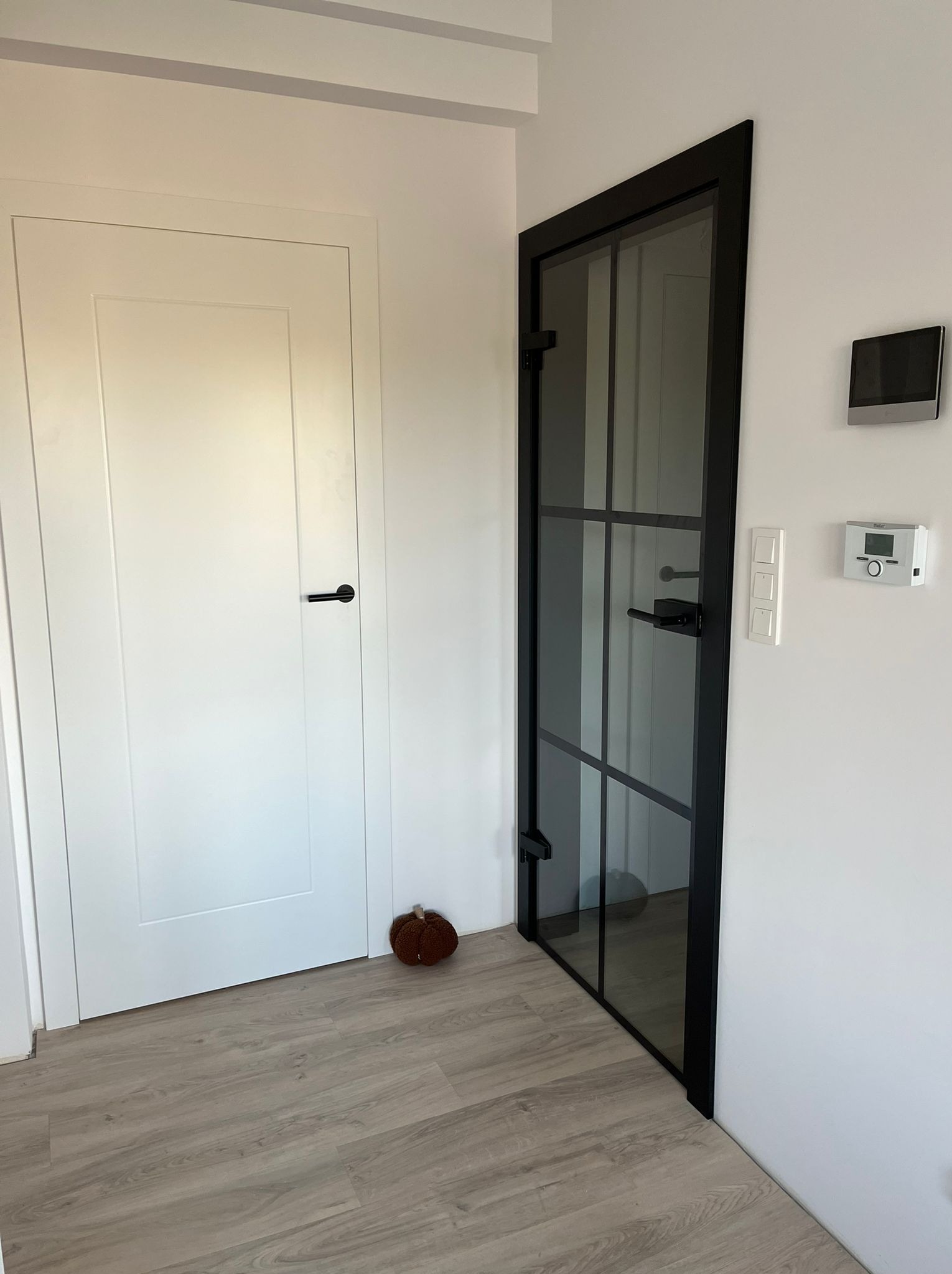 Wnętrze z dwiema drzwiami: białe drzwi z czarną klamką i czarne drzwi ze szprosami, na ścianie włączniki i termostat, na podłodze dekoracyjna dynia.