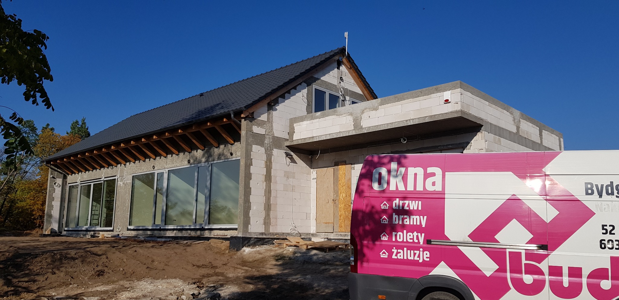 Budynek w trakcie budowy z zamontowanymi oknami, widoczny samochód firmowy z reklamą okien, drzwi, bram, rolet i żaluzji.