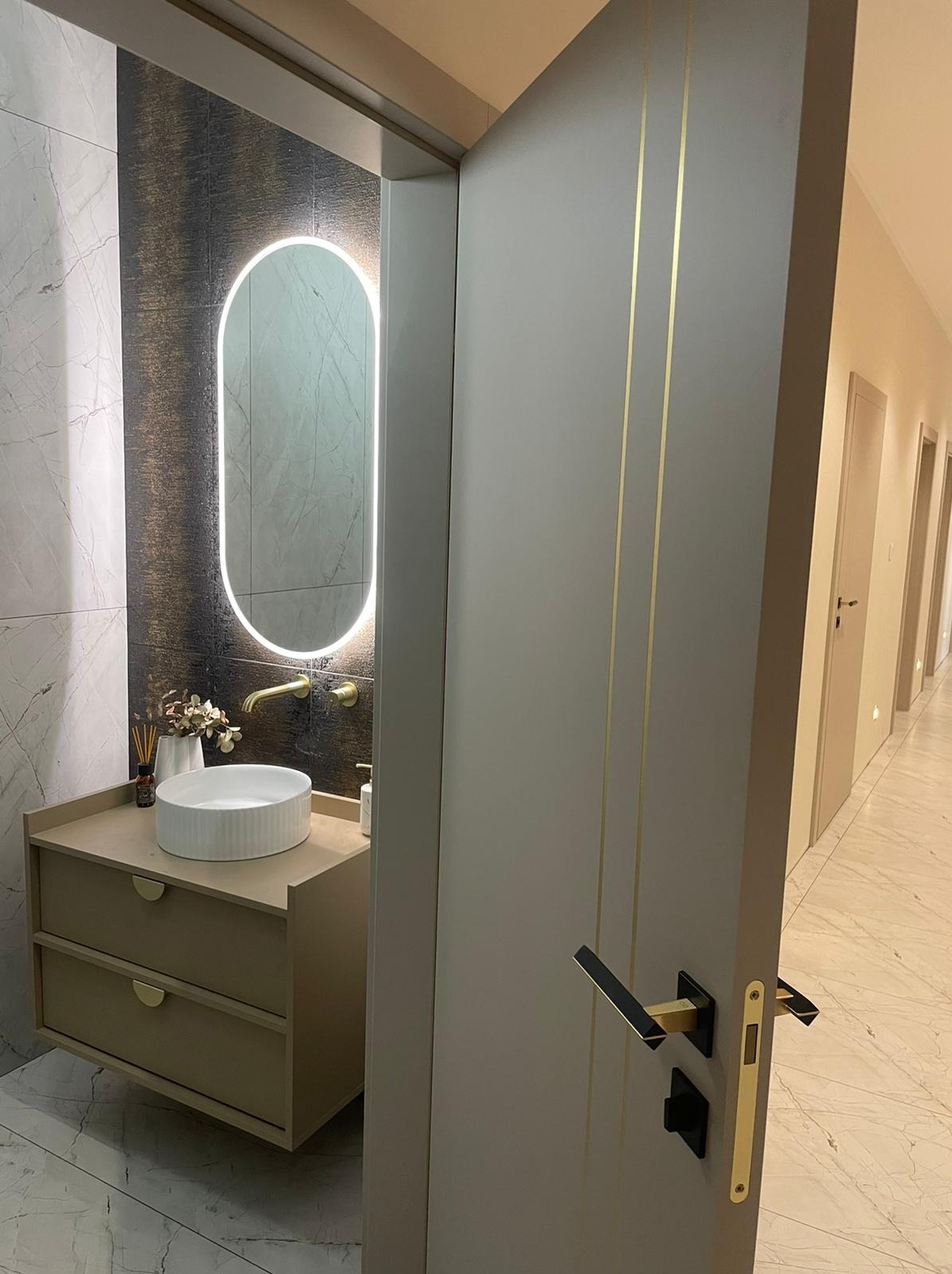 Otwarta szara drzwi z dwoma złotymi paskami pionowymi, widok na wnętrze łazienki z podświetlanym owalnym lustrem, umywalką na szafce z szufladami i marmurowymi płytkami na ścianach.