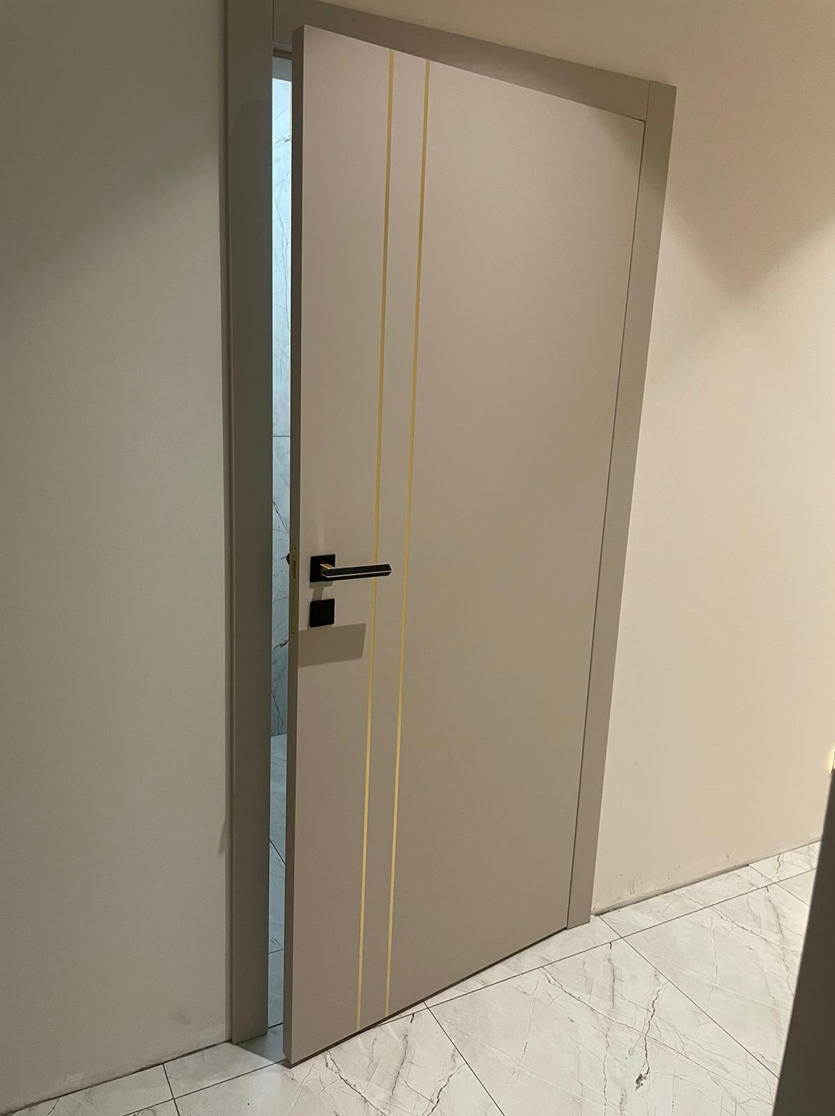 Beżowe drzwi wewnętrzne z dwoma pionowymi złotymi paskami, czarna klamka, widok na marmurową podłogę i częściowo otwarte pomieszczenie w tle.