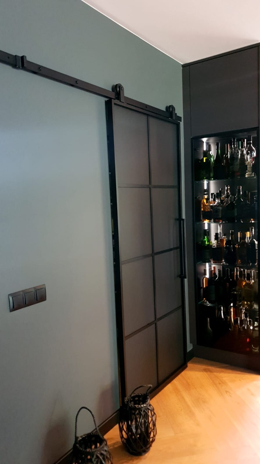 Ciemna, przesuwna, wewnętrzna brama z metalową ramą i podziałami, zamontowana na szynie, widoczna na tle ściany i podświetlonej szafki z alkoholem, z dwoma dekoracyjnymi lampionami na podłodze.