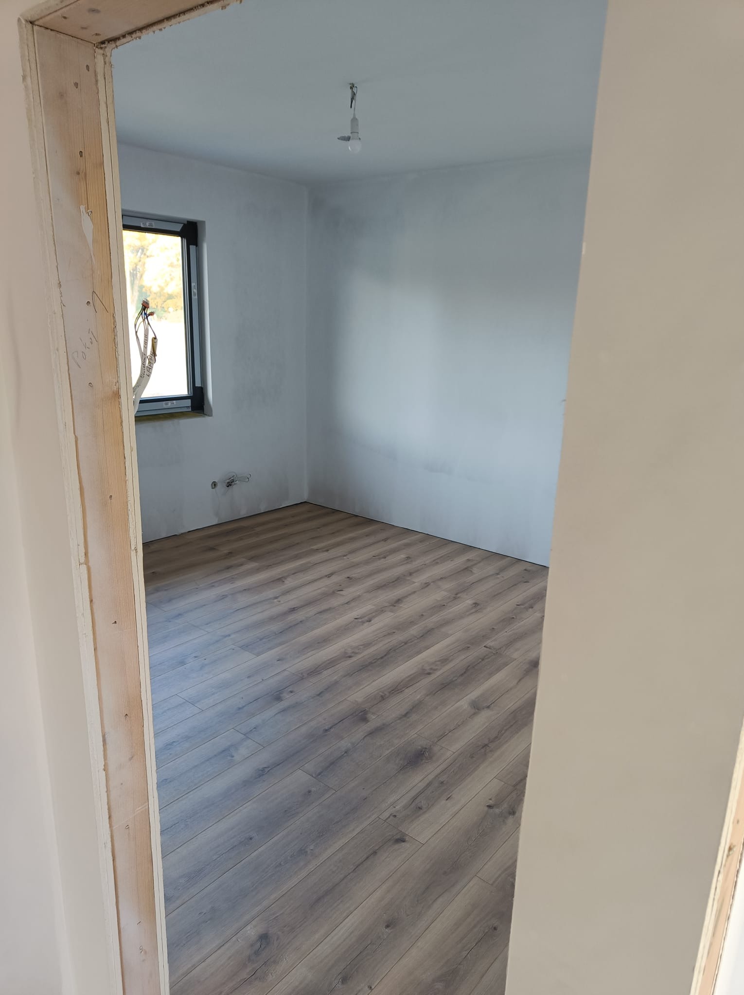 Wnętrze pokoju z panelami podłogowymi w odcieniach szarości, widok przez surową ościeżnicę, z widocznym oknem i instalacją elektryczną.