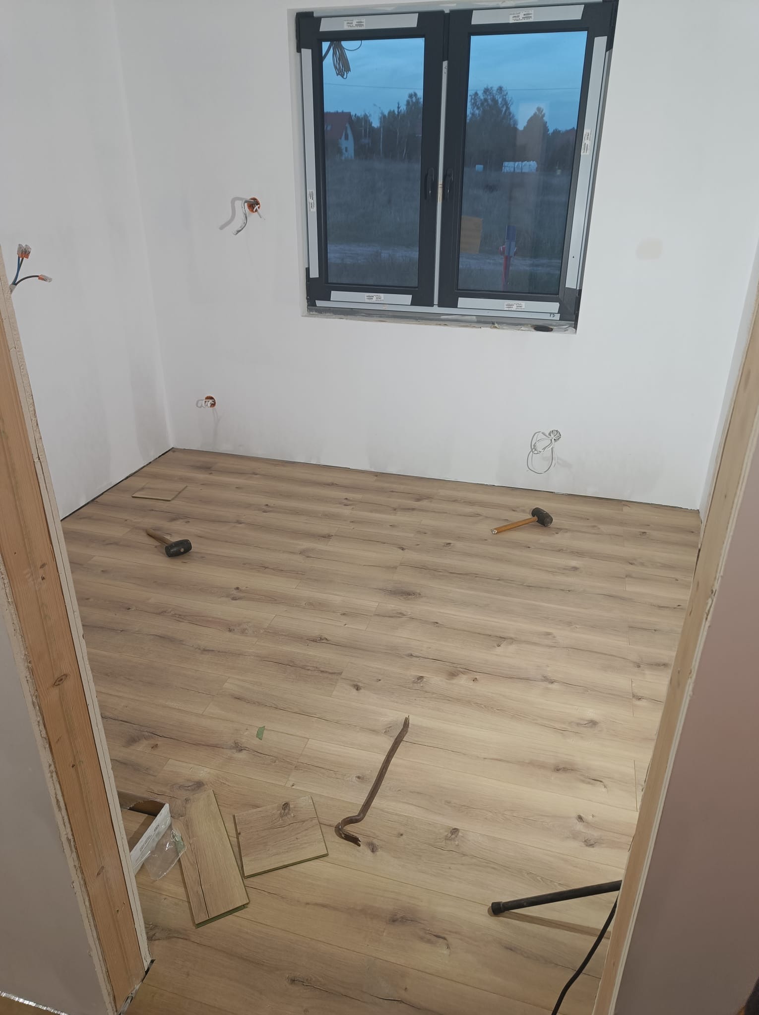Wnętrze pokoju w trakcie układania paneli podłogowych, widoczne narzędzia: młotek, łom, paczka paneli, okno w czarnej ramie.