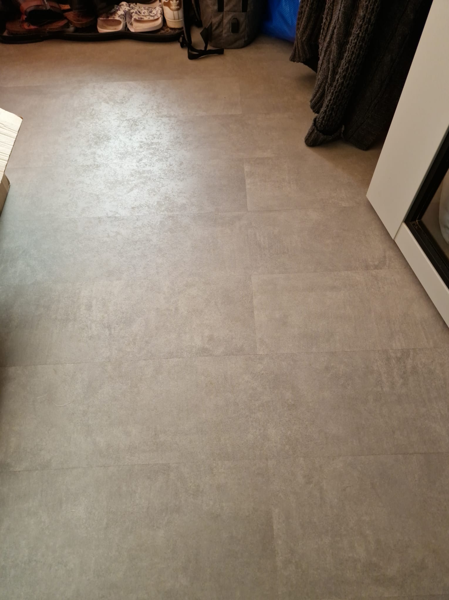 Szare panele podłogowe imitujące beton, ułożone w rzędach, widoczne buty i plecak w tle.