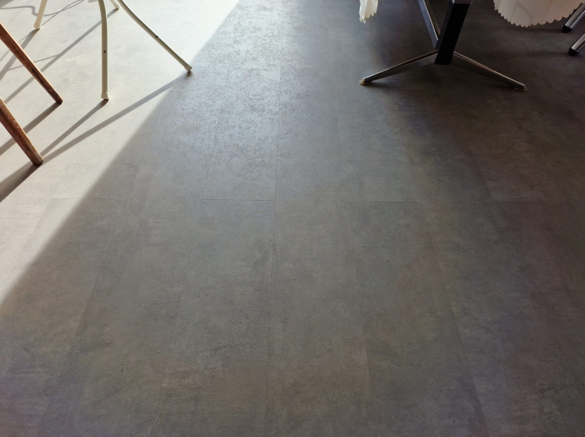 Szare panele podłogowe ułożone w regularny wzór, oświetlone naturalnym światłem z widocznymi nogami krzeseł i stołu.