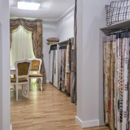 Wnętrze pracowni krawieckiej: próbniki tkanin wiszą obok stołu z krzesłami, w tle okno z dekoracyjną zasłoną i poduszkami. Drewniana podłoga.