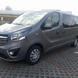 Szary Opel Vivaro, widok z przodu, zaparkowany na kostce brukowej, z budynkiem w tle. Auto z przyciemnianymi szybami, gotowe do transportu osób.
