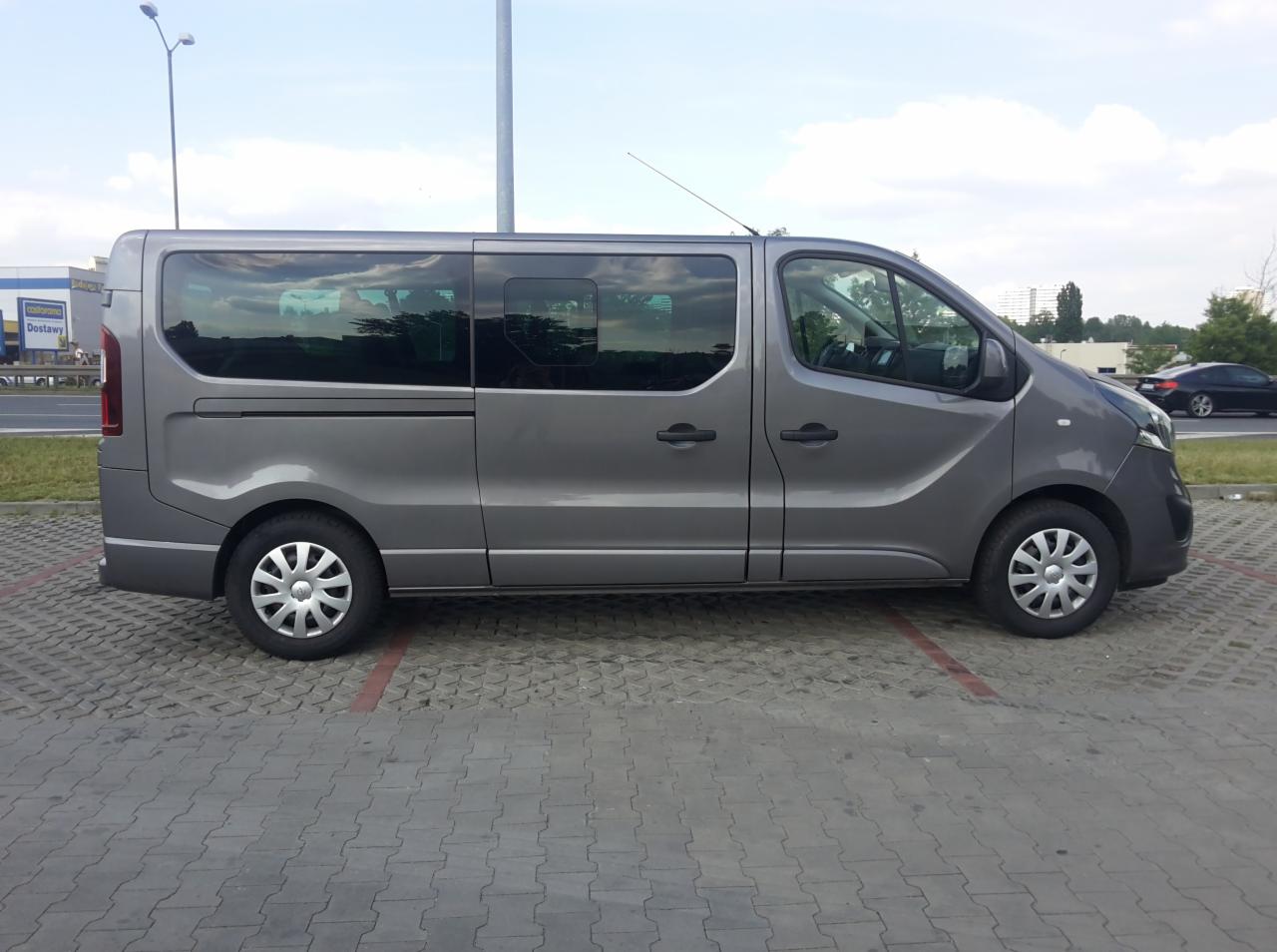 Szary, przeszklony van Renault Trafic na parkingu z kostki, widok z boku, z anteną na dachu. Tło: budynek sklepu i błękitne niebo z chmurami.