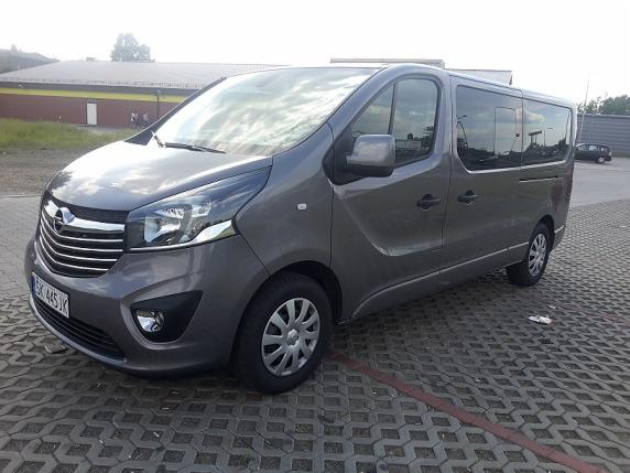 Szary Opel Vivaro, widok z przodu, zaparkowany na kostce brukowej, z budynkiem w tle. Auto z przyciemnianymi szybami, gotowe do transportu osób.