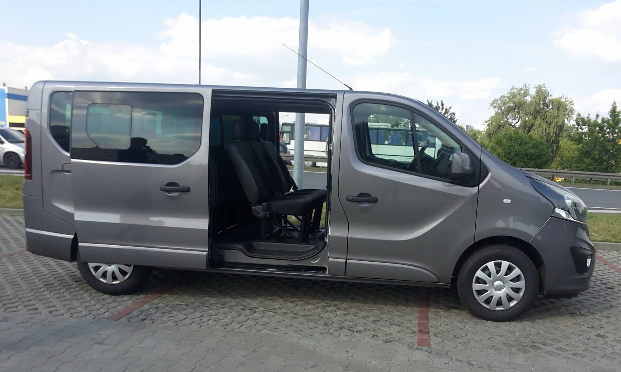Szary van z otwartymi drzwiami przesuwnymi, widoczne fotele pasażerskie z pasami bezpieczeństwa, zaparkowany na kostce brukowej przy drodze.