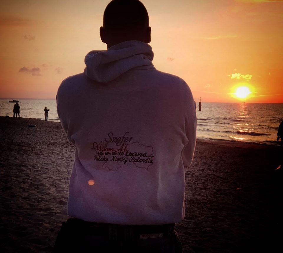 Mężczyzna w bluzie z logo firmy transportowej na tle zachodzącego słońca nad morzem.