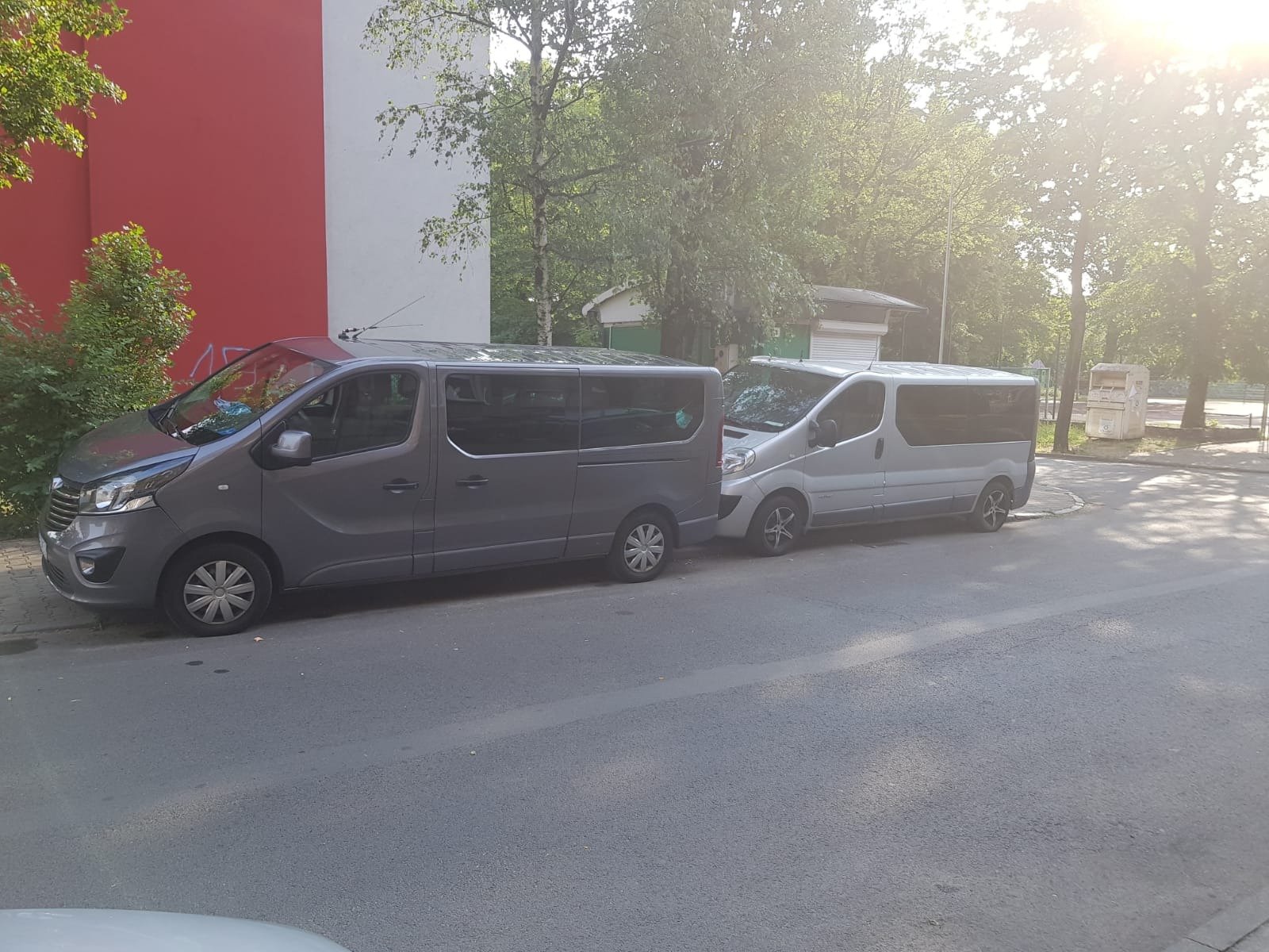 Dwa busy, szary i srebrny, zaparkowane obok siebie na ulicy w Chorzowie, przed budynkiem z czerwoną i białą ścianą, w tle zieleń drzew.