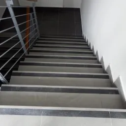 Klatka schodowa wyłożona szarymi płytkami z czarnymi antypoślizgowymi paskami, widok z góry na schody z metalową barierką, data wykonania zdjęcia w prawym dolnym rogu.