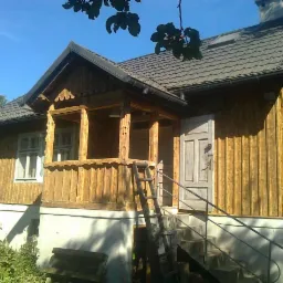 Drewniany dom jednorodzinny z poddaszem, drewnianą elewacją w pionowe deski, z werandą i schodami wejściowymi, po renowacji. Widoczne detale architektoniczne werandy.