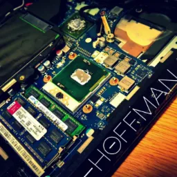 Otwarty laptop z widoczną płytą główną, procesorem, pamięcią RAM i elementami chłodzenia na drewnianym biurku. Napis 'HOFFMAN 4.0.4 BETA' na obudowie.