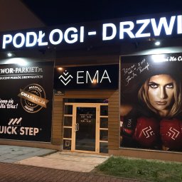 Podłogi Drzwi EMA Ardago - Fasada sklepu z podłogami i drzwiami, szyld z podświetlanym napisem 'PODŁOGI-DRZWI', reklamy producentów podłóg Jawor-Parkiet i Quick-Step, godziny otwarcia na drzwiach wejściowych i duży plakat...