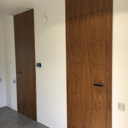 Podłogi Drzwi EMA Ardago - Dwa ukryte, fornirowane skrzydła w kolorze orzecha amerykańskiego, wkomponowane w białą ścianę, z minimalistycznymi, prostokątnymi klamkami i widocznymi włącznikami światła.