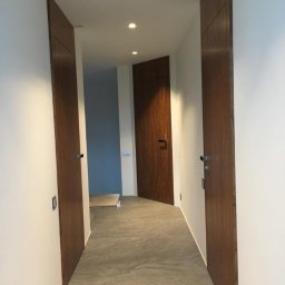 Podłogi Drzwi EMA Ardago - Korytarz z trzema drewnianymi drzwiami o minimalistycznym designie i szarą podłogą z płytek imitujących kamień, oświetlony punktowymi lampami sufitowymi.