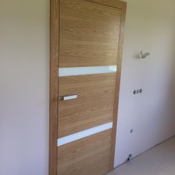 Podłogi Drzwi EMA Ardago - Zamknięte drewniane drzwi wewnętrzne z dwoma poziomymi paskami z matowego szkła, zamontowane w jasnym pomieszczeniu z widocznymi instalacjami na ścianie.