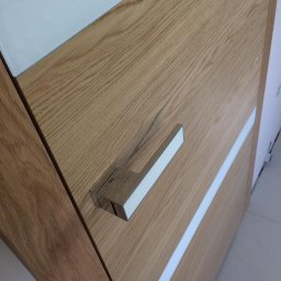 Podłogi Drzwi EMA Ardago - Fragment drewnianej powierzchni z chromowaną klamką i białym paskiem dekoracyjnym, widoczne usłojenie drewna.