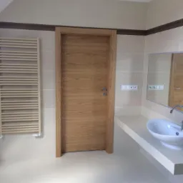 Jasna łazienka z drewnianą okleiną na drzwiach, widoczny grzejnik drabinkowy i umywalka nablatowa.