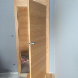 Podłogi Drzwi EMA Ardago - Montaż dębowych drzwi wewnętrznych z poziomym usłojeniem i srebrną klamką, widoczne ślady kleju do parkietu na podłodze, szare ściany.