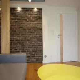 Białe drzwi wewnętrzne z pionowym, jasnym paskiem w nowoczesnym salonie z ceglaną ścianą i drewnianym słupem, widoczne fragment sofy i żółtego stolika.