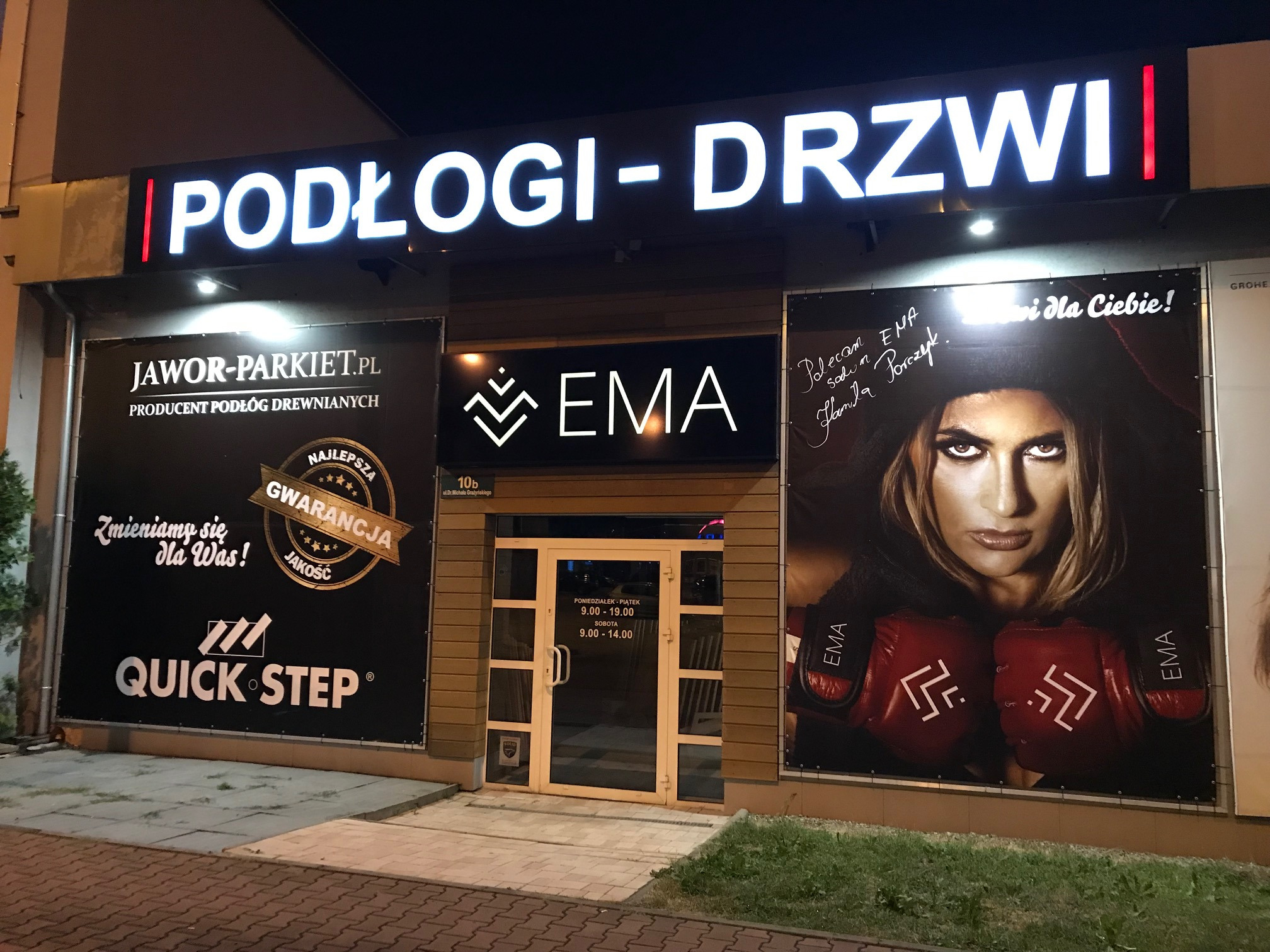 Fasada sklepu z podłogami i drzwiami, szyld z podświetlanym napisem 'PODŁOGI-DRZWI', reklamy producentów podłóg Jawor-Parkiet i Quick-Step, godziny otwarcia na drzwiach wejściowych i duży plakat...