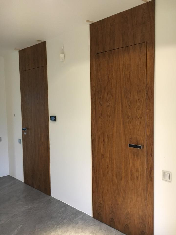 Dwa ukryte, fornirowane skrzydła w kolorze orzecha amerykańskiego, wkomponowane w białą ścianę, z minimalistycznymi, prostokątnymi klamkami i widocznymi włącznikami światła.