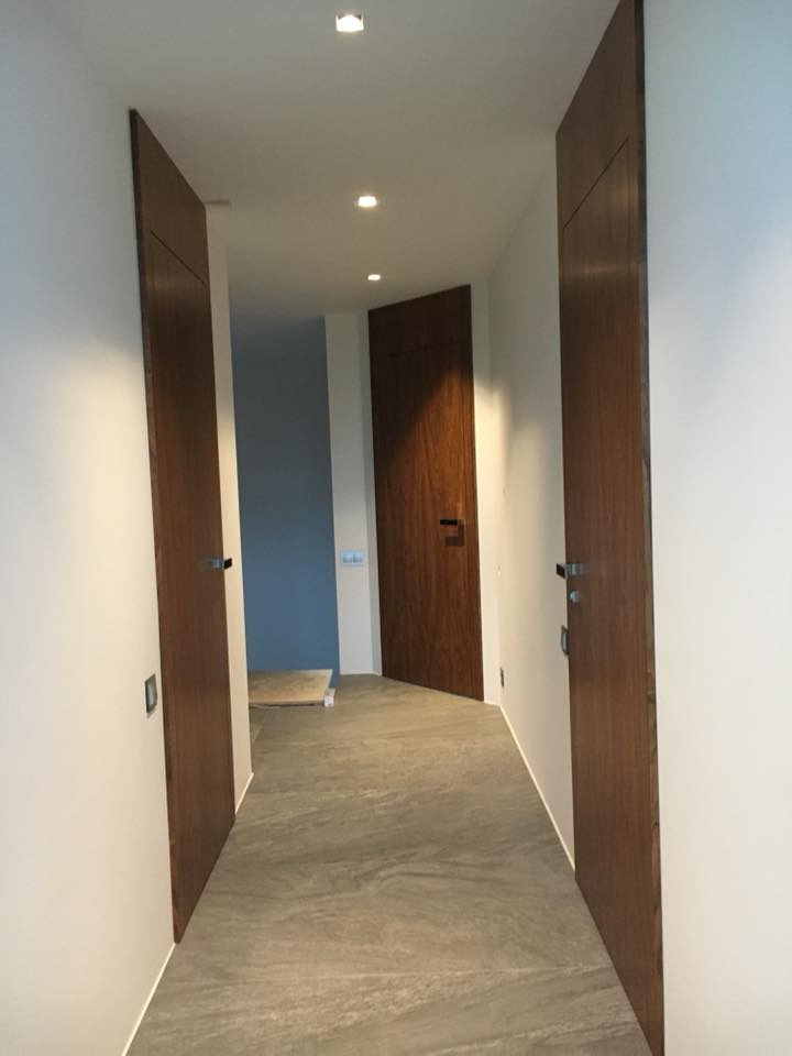 Korytarz z trzema drewnianymi drzwiami o minimalistycznym designie i szarą podłogą z płytek imitujących kamień, oświetlony punktowymi lampami sufitowymi.