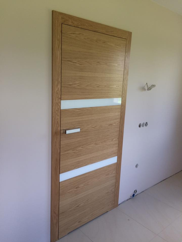 Zamknięte drewniane drzwi wewnętrzne z dwoma poziomymi paskami z matowego szkła, zamontowane w jasnym pomieszczeniu z widocznymi instalacjami na ścianie.