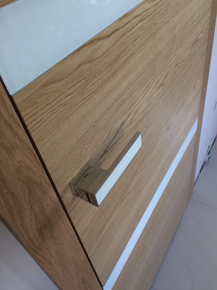 Fragment drewnianej powierzchni z chromowaną klamką i białym paskiem dekoracyjnym, widoczne usłojenie drewna.