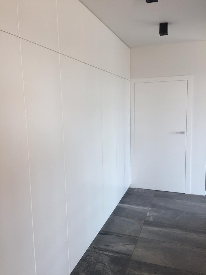 Minimalistyczne wnętrze: Biała ściana z ukrytymi szafami, białe drzwi z prostą klamką, szara podłoga z płytek imitujących kamień, czarne reflektory sufitowe.