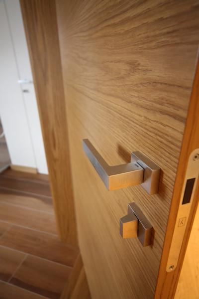 Częściowy widok skrzydła drzwiowego z okleiną dębową i nowoczesną, prostokątną klamką w kolorze satynowego niklu, widoczna podłoga z paneli drewnianych w odcieniach brązu.