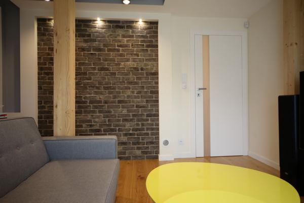 Białe drzwi wewnętrzne z pionowym, jasnym paskiem w nowoczesnym salonie z ceglaną ścianą i drewnianym słupem, widoczne fragment sofy i żółtego stolika.