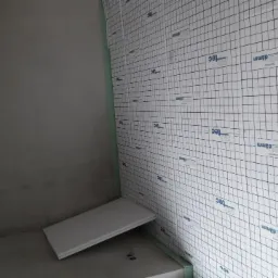 Wnętrze w trakcie prac przygotowawczych do wykończenia, ściana wyłożona płytami termoizolacyjnymi z nadrukowaną siatką i logo producenta, surowa ściana z betonu obok.