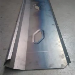 Metalowa osłona z wyciętymi otworami w kształcie trapezu, uformowana z blachy, leżąca na betonowej posadzce. Widoczne ślady obróbki i punktowe przebarwienia termiczne.