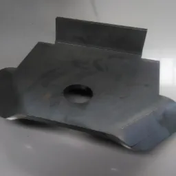 Szara, metalowa płyta z otworem i przyspawanym prostopadłościanem, leżąca na jasnej powierzchni.