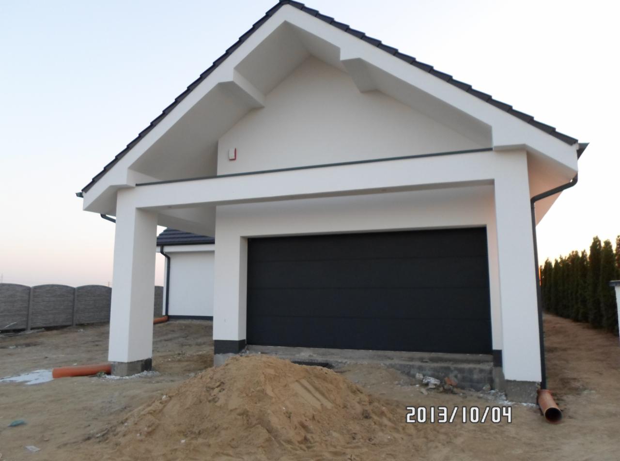 Nowoczesny garaż z czarną bramą segmentową, białe ściany i filary, widoczny fragment dachu i otoczenia. Ujęcie z poziomu gruntu, data w prawym dolnym rogu.